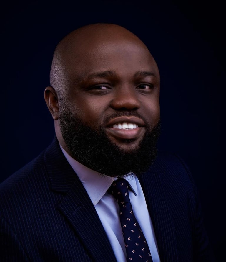 Professional headshot of a man with dark skin, full beard, and shaved head wearing a navy pinstripe suit, light blue shirt, and dark patterned tie against a dark background