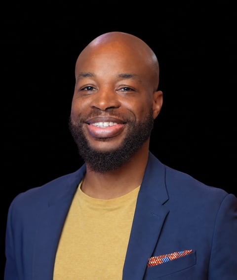 Professional headshot of a smiling man with a beard wearing a navy blazer over a yellow shirt against a black background