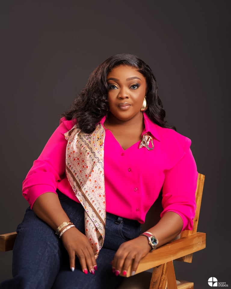 Professional portrait of a woman in a bright pink blouse and dark jeans seated on a wooden chair against a gray background