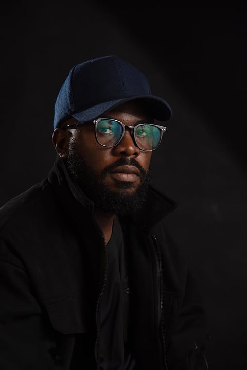 Portrait of a man wearing a blue baseball cap, clear-framed glasses, and black jacket against a dark background