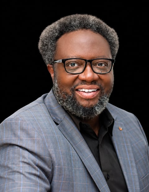Professional headshot of a man with gray hair and beard, wearing glasses and a light blue plaid blazer over a dark shirt, smiling against a black background.